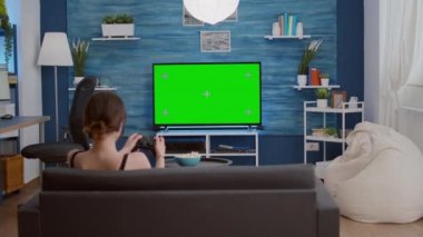 Oyuncu kız yeşil ekran TV 'de kablosuz kontrolörle video oyunu oynuyor. Kanepede dinleniyor.