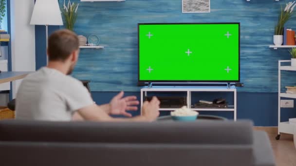 Homem sentado no sofá assistindo jogo de esporte na tela verde tv mockup incentivando a equipe favorita — Vídeo de Stock