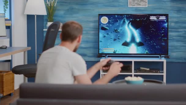 Hombre sosteniendo el controlador inalámbrico jugando videojuego de consola tirador en primera persona en la televisión mientras está sentado en el sofá — Vídeo de stock