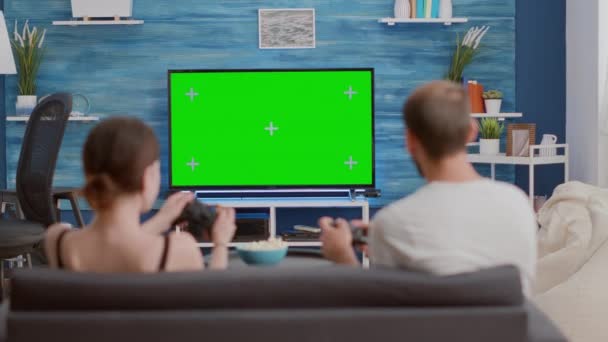 若い女性とボーイフレンドゲーマーのバックビューは、コントローラーを保持している緑色の画面テレビでコンソールでアクションゲームをプレイ — ストック動画