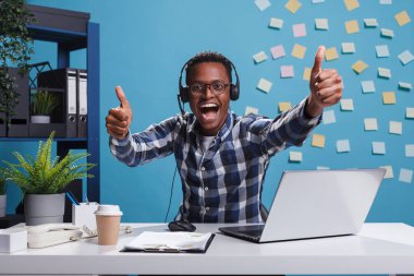 Aşırı mutlu gülümseyen çağrı merkezi operatörü kameraya bakarken parmaklarını kaldırıyor..