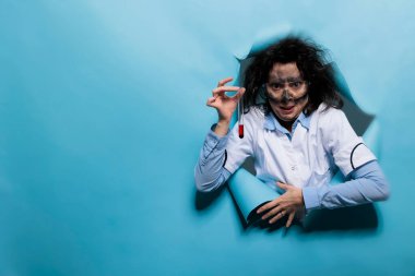 Dağınık saçlı, pis sırıtan ürkünç suratlı çılgın biyokimya uzmanı cam test tüpü kimyasal karışımla doluyken. Mavi arka planda şapşal görünüşlü çılgın bir kimyager. Stüdyo çekimi