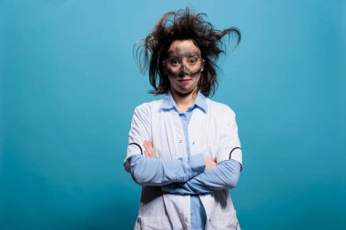 Komik görünümlü, dağınık saç stili ve kirli yüzlü çılgın bir kimyager mavi arka planda kameraya bakıyor. Düzensiz saçları ve kirli yüzüyle kollarını kavuşturmuş komik çılgın bilim adamı..