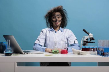 Laboratuvar patlamasından sonra masasında oturan dağınık saçlı ve kirli yüzlü aptal görünümlü kimyager. Mavi arka planda oturan çılgın görünümlü aptal bir laboratuvar çalışanı. Stüdyo çekimi