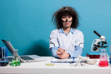 Crazy mad biochemistry expert with messy hair and dirty face sitting at lab desk after experiment explosion. Maniac goofy looking chemist looking dumb while looking at camera on blue background.