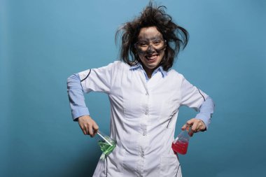 Erlenmeyer şişeleri sıvı maddelerle doluyken tuhaf sırıtan çılgın biyokimya uzmanı. Kirli yüzlü, dağınık saçlı, çılgın görünüşlü bir kimyager mavi arka planda aptalca davranıyor.