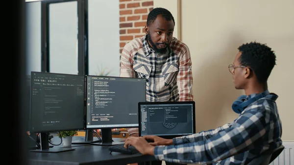 Software developer sitting at desk with multiple screens and laptop running code talking with colleague about artificial intelligence algorithm. Software developers doing teamwork in it office.