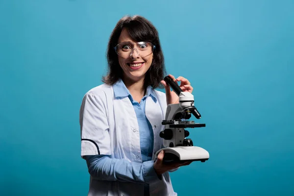 Insane looking scientist smiling creepy while having modern microscope instrument on blue background. Lunatic chemist with gruesome smile having electronic magnifying laboratory instrument.