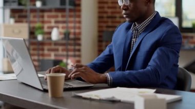 Afrikalı Amerikalı düşünceli bir adam dizüstü bilgisayarla proje planlamak için beyin fırtınası yapıyor. Düşünceli yönetici web sitesindeki sunum raporunu düşünüyor, araştırma e-postası yazıyor.