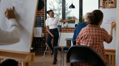 Sanat sınıfı atölyesindeki öğrencilere çizim becerilerini açıklayan genç öğretmen. Kadın sanatçı insanlara beyaz tuval ve ahşap sehpaya vazo çizmeyi öğreterek kişisel gelişime yardımcı oluyor..