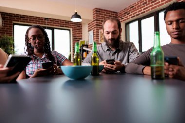 Modern akıllı telefon cihazları olan çok kültürlü arkadaş grubu sosyal medyada gezinirken evde oturuyorlar. Oturma odasındaki çeşitli insanların cep telefonları internette sörf yapıyor..