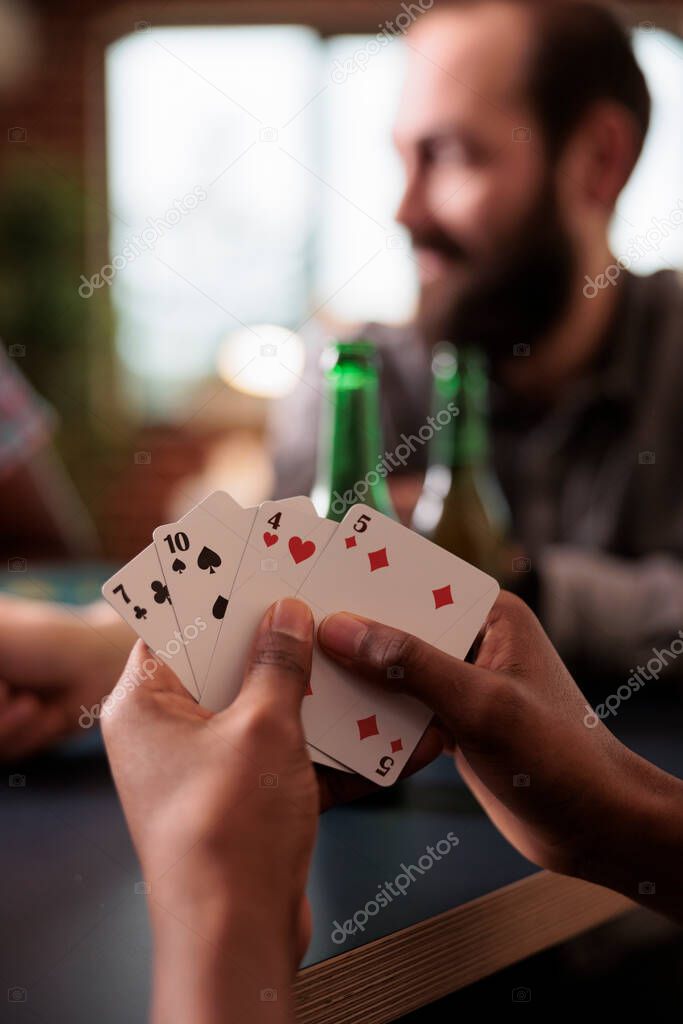 Close up shot of african american hands holding cards while playing with friends at home. Diverse group of people sitting at home in living room while relaxing with snacks, beer and society games.