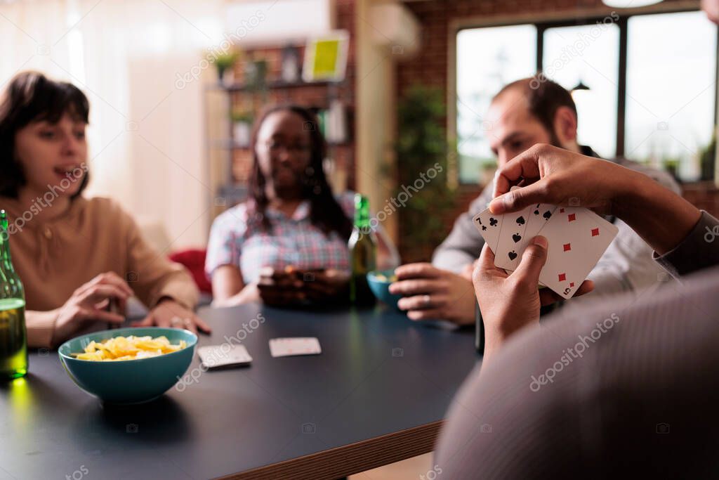 Selective focus of african american man holding cards while playing with best friends at home. Happy multiethnic people sitting at table in living room while enjoying fun leisure activity together.