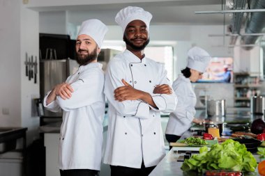 Mutfak alanındaki kameralara bakarken yemek endüstrisi çalışanları sırt sırta veriyor. Yemek servisi için taze malzemeler hazırlarken kolları çapraz duran kendinden emin aşçılar..