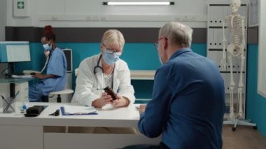 Genel pratisyen ve yüz maskeli hasta tıp bürosunda ilaç şişesinden bahsediyorlar. Hekim, hastalıkları tedavi etmek için emekli adama reçeteli tedavi ve ilaç veriyor..