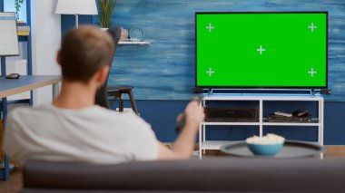 Static tripod shot of man switching channels while looking at green screen on tv and sitting on sofa. Over shoulder view of person relaxing using television remote zapping on chroma key display. clipart
