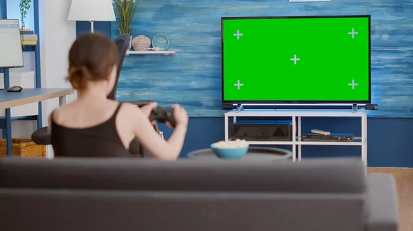 Static tripod shot of gamer girl holding wireless gamepad playing action console video game on green screen tv while sitting on sofa. Woman enjoying online gaming on chroma key display with controller