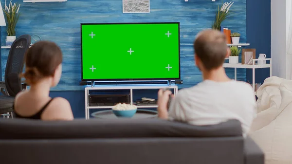 Back view of young woman asking boyfriend to change tv channel on green screen while spending time on couch. Couple enjoying television shows on chroma key mockup screen in modern living room.