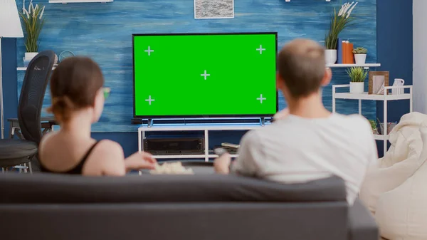 Couple sharing a bowl of popcorn while watching tv and switching channels looking at green screen in modern living room. Woman and man watching chroma key television and zapping while having a snack.