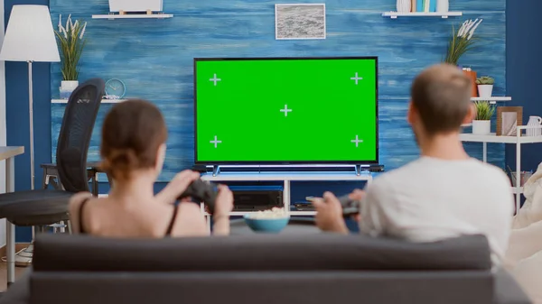 Back view of young woman and boyfriend gamers holding controllers playing action game on console on green screen tv sitting on couch. Couple spending free time gaming on chroma key mockup display.
