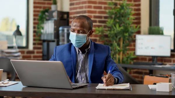 African american worker planning business statistics on papers and laptop network, using data report to take notes. Office employee doing online paperwork on website browser and files.