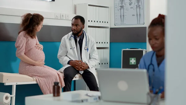 Patient expecting child meeting with physician in medical cabinet, attending exam appointment to receive pregnancy advice. Pregnant woman talking to male doctor about childbirth.