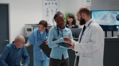 Afro-Amerikalı kadın ve doktor, dijital tablet üzerindeki teşhisi sağlık desteği için analiz ediyorlar. Bekleme odasının lobisinde, hastane resepsiyonunda, check-up muayenesinde çeşitli insanlar konuşuyor..