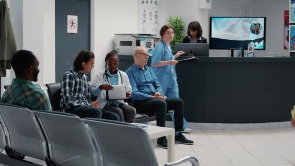 アフリカ系アメリカ人医師が待機地域でアジア人患者と話をし 医療スタッフと病院の受付ロビーで相談を行う 待合室でおしゃべりする多様な人々 — ストック動画