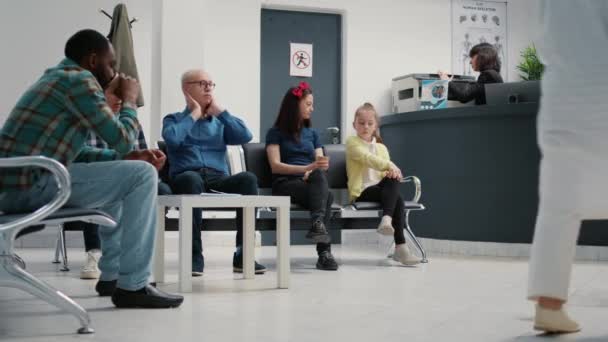 病院の受付で待合室に座っている患者の多くは 予約で視察を開始する準備をしています 待合室で子供 高齢者や他の人と母 三脚ショット — ストック動画