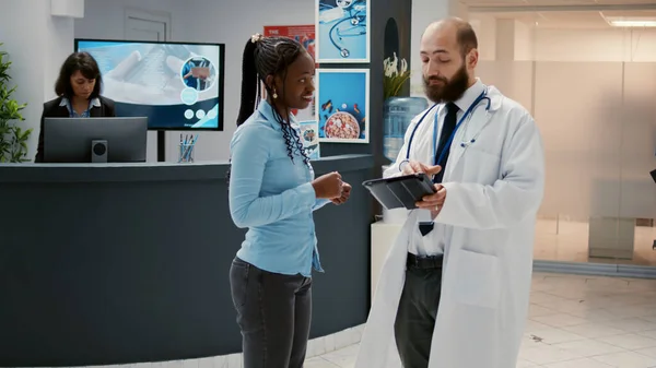 男医生与非洲裔美国病人谈论医疗保健 在医院接待处候诊室大厅的数字平板电脑上显示医疗诊断结果 设施的检查访问 — 图库照片