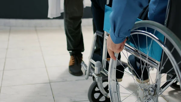 Senior patient with chronic disability using wheelchair in waiting room at hospital reception area to meet with doctor for appointment. Man with health condition doing checkup visit. Close up.