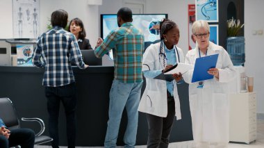 Muhtelif tıbbi ekipler bekleme alanındaki kontrol raporlarını inceliyor, danışma hakkında konuşuyorlar. Doktorlar sağlık hizmetlerini tartışıyor, hastane resepsiyonundaki belgelere bakıyorlar..