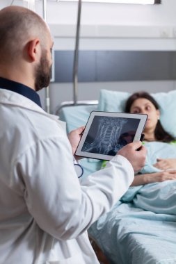 Uzman doktor, hastane koğuşunda çalışan tablet bilgisayarla kemik radyografisini açıklayan hasta kadını izliyor. Hasta, hastalık sonrası yatağında dinleniyor.