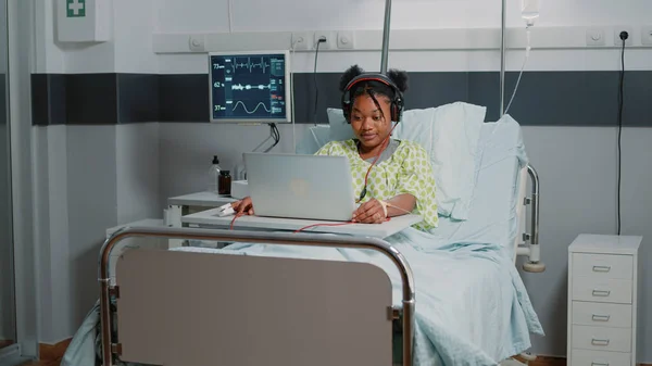 Patient with sickness using headphones to listen to music while sitting in hospital ward bed. Young woman looking at laptop and listening to audio sounds on headset. Adult with disease