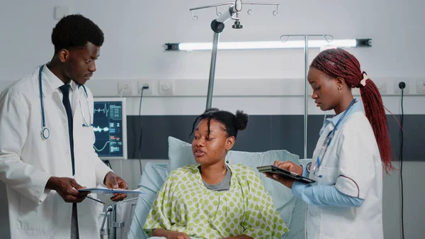 Team of african american doctors consulting sick patient in bed. Medical specialists discussing about healthcare treatment and medicine to cure illness while woman holding tablet.