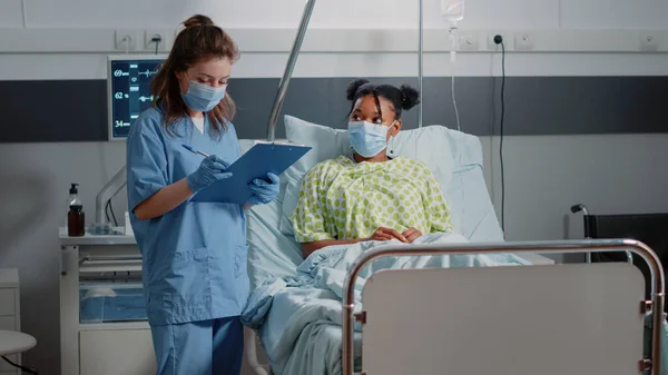 病棟でフェイスマスクを着用し 病気の患者と相談を行う医療アシスタント 保健医療について若い女性と話し合い パンデミックの際に注意を払う看護師 — ストック写真