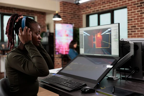 Game artist in office workspace sitting at desk while working on game interface. Level design department employee wearing wireless headphones while analyzing digital environment