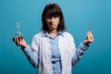 Laboratuvarda çalışan bir kimyager kameraya bakarken içi sıvı maddeyle dolu cam şişeler tutuyor. Bilinmeyen bileşiklerle dolu cam şişeler tutan kendinden emin bir araştırma uzmanı..