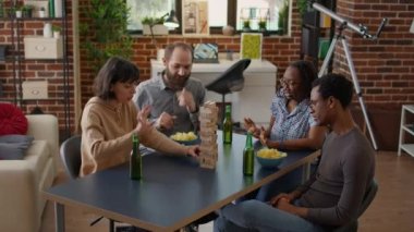 Competitive woman crashing blocks tower and losing game, laughing with friends at board games gathering. People having fun with wooden construction cubes and square pieces at home.