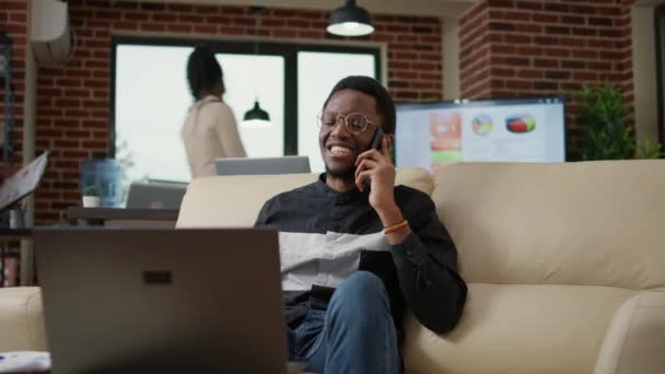 非裔美国人在商务办公室打电话聊天 用智能手机享受与同事的远程通信 在笔记本电脑上工作和谈话的年轻人 — 图库视频影像