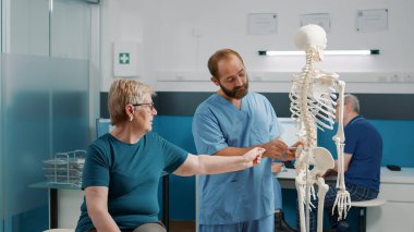 Osteopat, insan iskeleti üzerindeki ortopedik teşhisi iyileşme kontrolündeki yaşlı kadına açıklıyor. Sağlık uzmanı fiziksel yaralanma hakkında konuşmak için omurilik ve anatomi kemiklerine işaret ediyor..