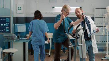 Kıdemli hasta kaslı bisiklet üzerinde kas egzersizi yapıyor kas iskeleti ve mekanik bozuklukları tedavi etmek için. Fizyoterapi bisikleti kullanan yaşlı kadına tıbbi danışmanlık yapıyor..
