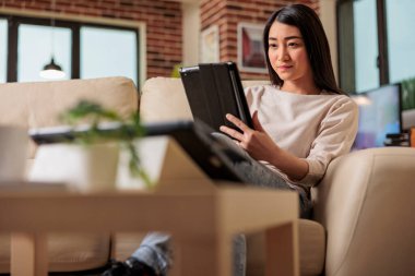 Evinde dijital tablet kullanan genç ve güzel Çinli bir kadın kanepe eğlence cihazının üzerinde otururken internet ağı 3.0 'ı bağlarken gülümsüyor, kablosuz bilgisayar tableti kullanıyor.