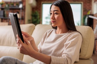 Evinde dijital tablet kullanan genç ve güzel Çinli bir kadın kanepe eğlence cihazının üzerinde otururken internet ağı 3.0 'ı bağlarken gülümsüyor, kablosuz bilgisayar tableti kullanıyor.