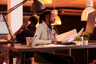 Serbest çalışan kadın, iş evraklarını veri tablolarıyla analiz ediyor, bilgisayarda sunum yapıyor. Gün batımında şirket raporu üzerinde çalışmak için araştırma bilgilerine sahip belgelere bakıyorum..