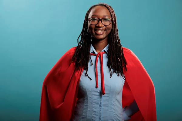 Smiling heartily young adult superhero woman wearing red hero cape while posing at camera. Portrait of brave and proud african american woman being happy and joyful on blue background. Studio shot.