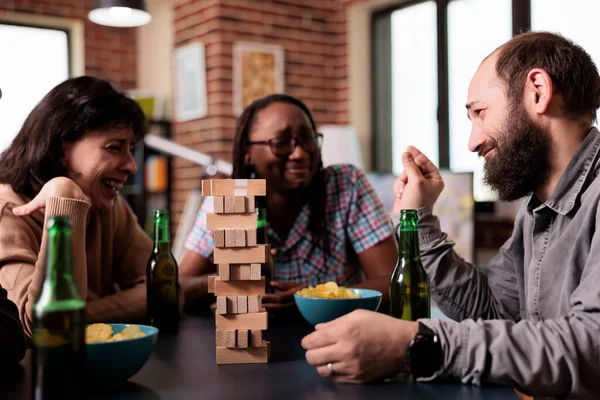 Laughing diverse people sitting at table while talking and enjoying society games together. Multiethnic people sitting at home in living room while discussing and playing with wood blocks.