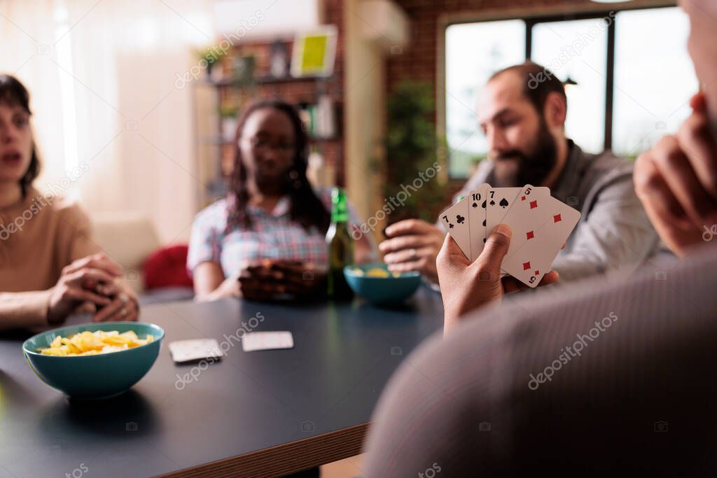 Selective focus of man holding cards in hand while playing with friends at home. Pensive person thinking about next move while playing society games and enjoying snacks and beverages.