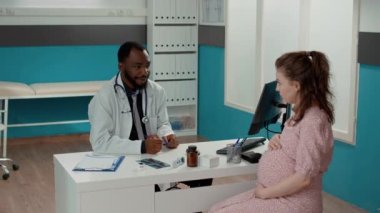 Genel pratisyen hekim kadın ofiste çocuk bekliyor, muayene randevusuna katılıyor. Erkek doktor hamile bir hastayla özel bir konuşma yapıyor..