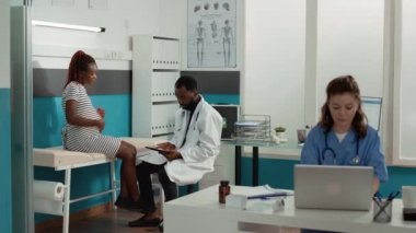 Hamile kadınlara danışmak için dijital tablete yazı yazan erkek doğum uzmanı, sağlık kontrolünde notlar alıyor. Doktor, çocuk bekleyen hastalara tıbbi danışmanlık yapıyor, doğum tavsiyesi veriyor..